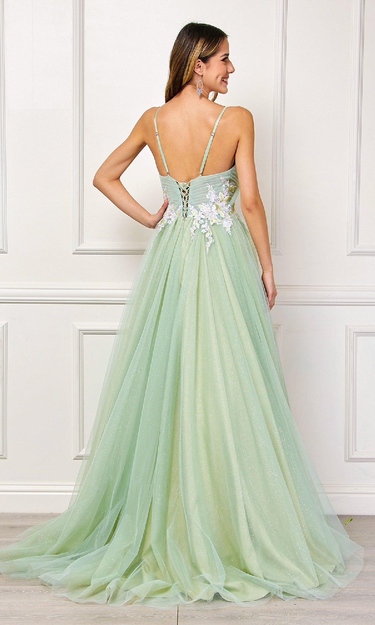 A woman stands before a white paneled wall wearing the Amelia Couture SU088, a long pastel prom ball gown with floral embroidery, lace-up back, and spaghetti straps.